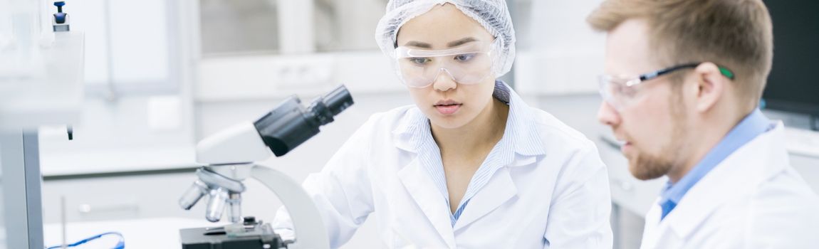 2 science students in a lab wearing protective clothing sitting beside a microcope