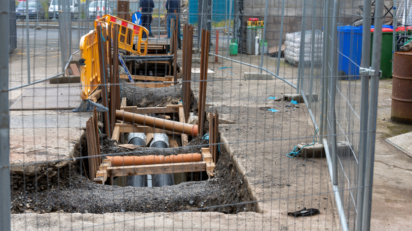 ground exposed in construction area to show large black pipes underneath. The area is fenced off.