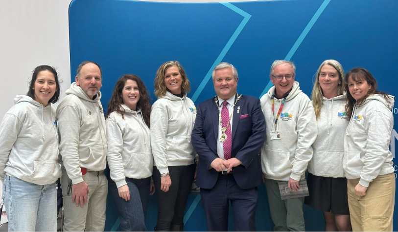 5 women and 3 men smile for camera. 7 in similar grey branded hoodie and one man in suit with medal of deputy lord mayor around his neck