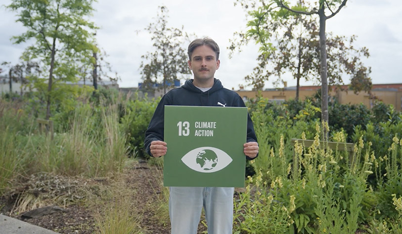 Young white man holding a square green sign with SDG13 Climate Action against backdrop of plants and foliage
