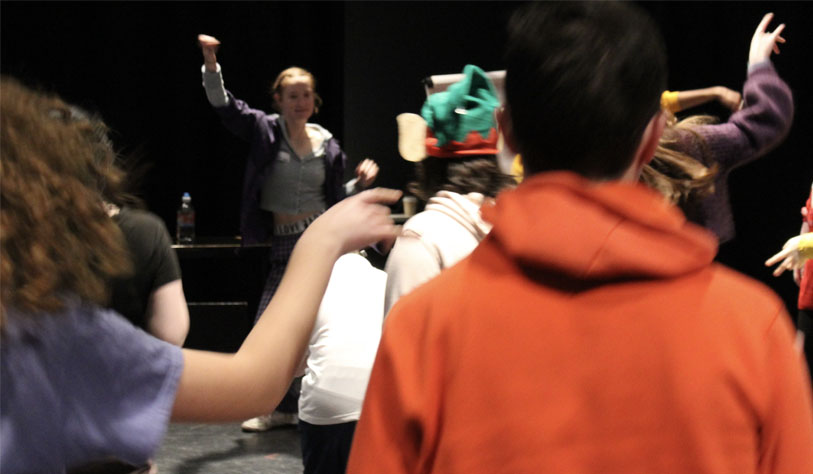 Young people dancing in a black stage set. some wear bright clothes that pop against the dark