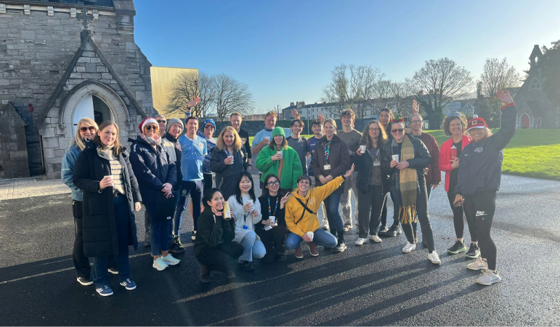 Group of around 30 people smile for camera against campus backdrop a grey building and blue sky