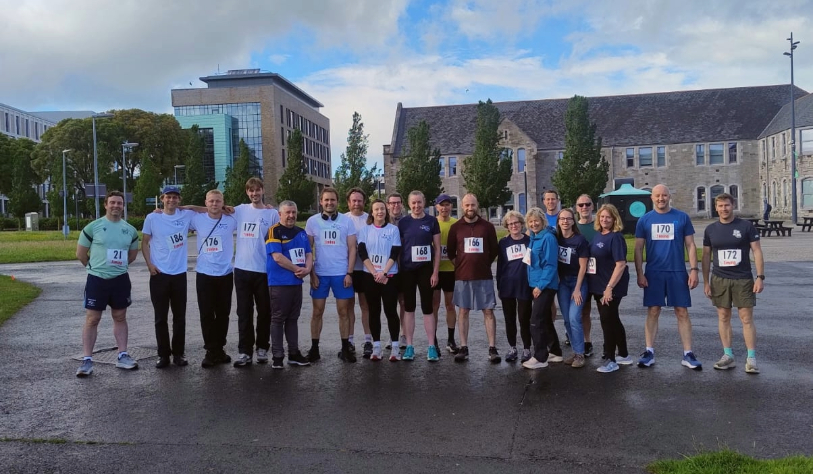 group of 20 people wearing running gear and race numbers pose for camera against campus backdrop with mixture of old and new buildings