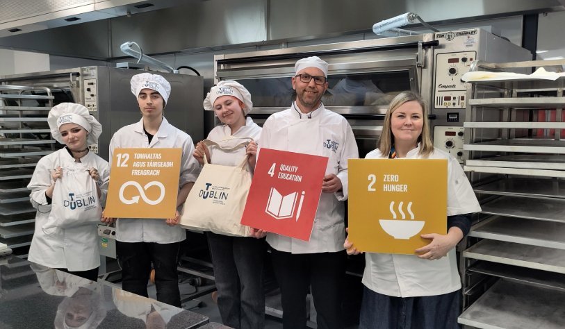 5 people in bakery uniform in backdrop of large ovens in kitchens. one holds bread bag, 3 holding Sustainable Development Goals signs