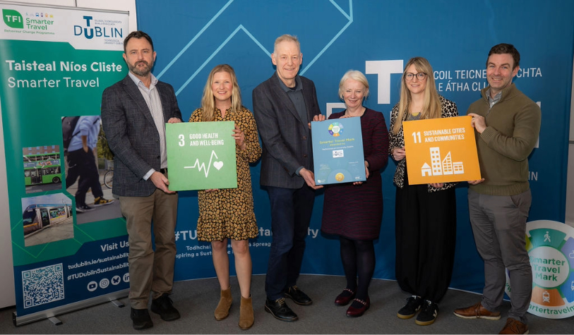 three male and three female hold plaque and sdg signs 11 and 3 against tu dublin backdrop and smile for camera