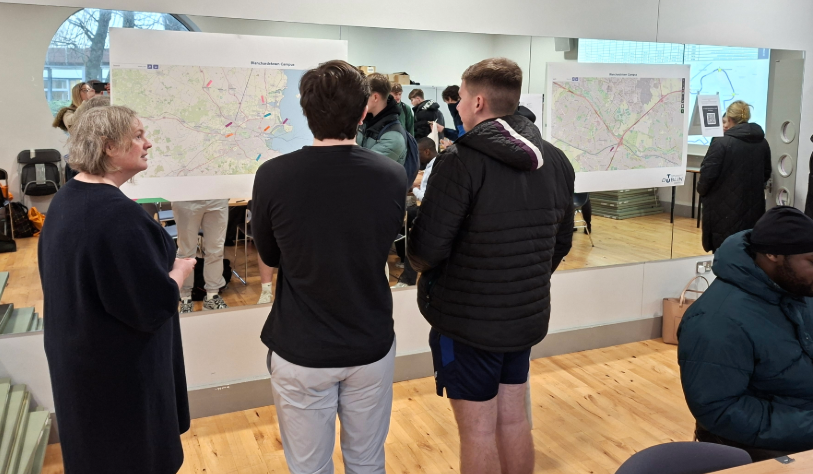 one woman and three young men stand in front of large map. they face away from us. there are others in the background which can be seen as one wall is mirrored