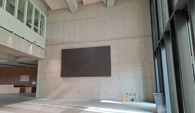 large high ceilinged room in bare concrete containing large digital screen on wall mid way up