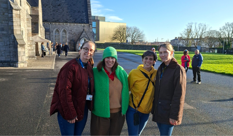 four women  smile for camera in campus backdrop of grey church building, blue sky and green grass