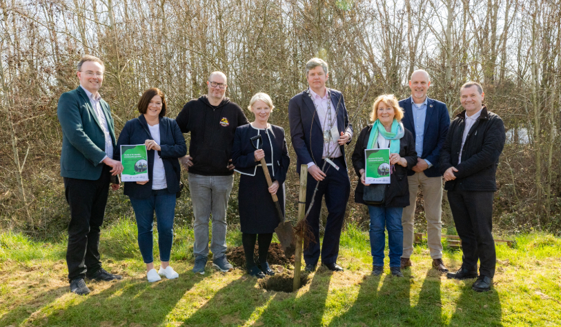 three white women and four white men smile for camera in outdoor setting. centre man holds shovel and centre woman holds tree