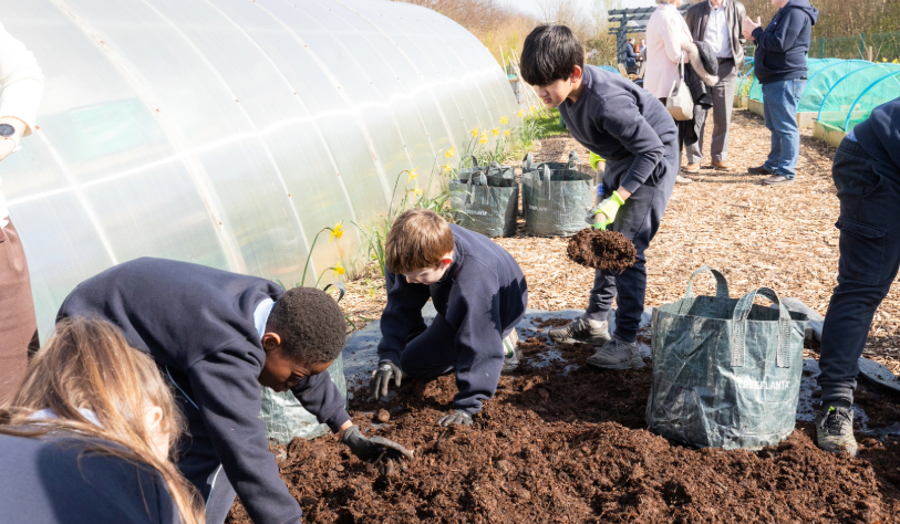 five school children shovelling soil, three older adults chatting in background