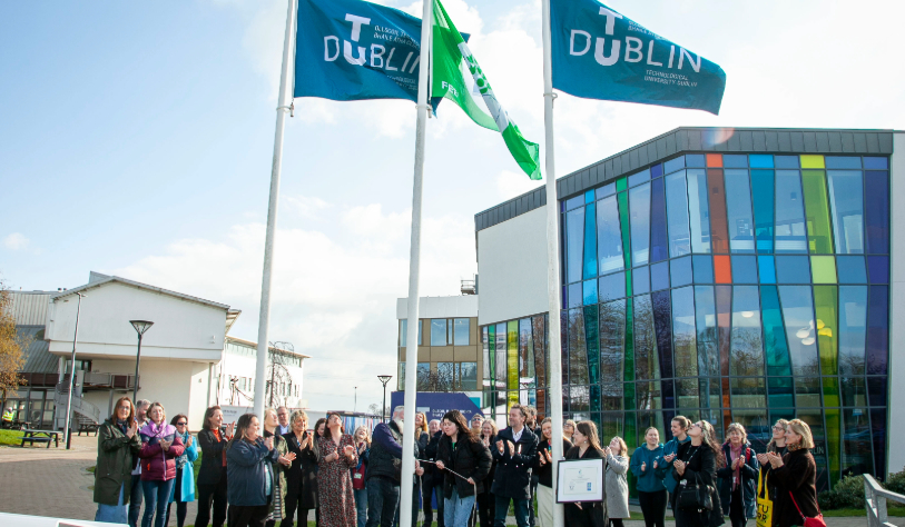 group of 40 plus people look up as Green flag is raised among two other TU Dublin flags against campus backdrop