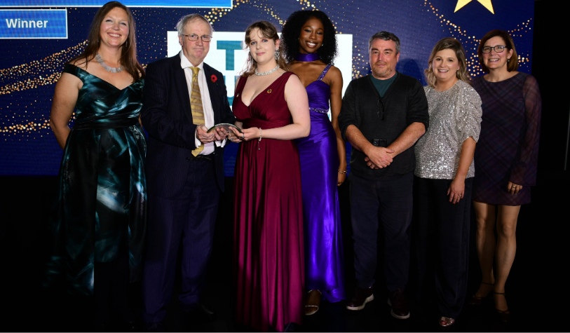 female student receives award from male. four other females and one other male also smile for camera against awards backdrop. most are in formal dress
