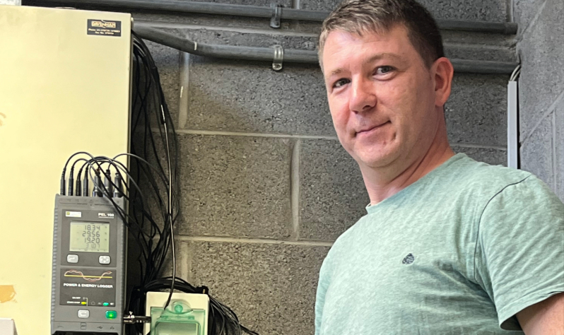white male looks to camera and stands beside smart energy meter fixed to wall
