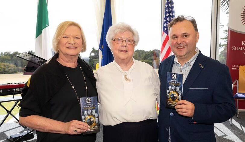 An image of three smiling people standing in front of an Irish, European and American flag