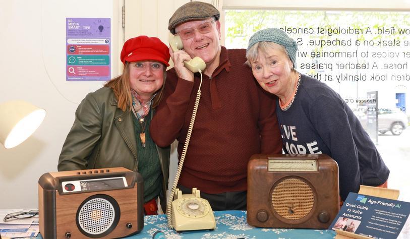Three people post in front of two old fashioned radios and a telephone