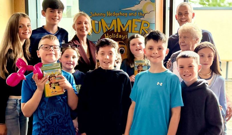 group of children and adults pose together indoors, with several children holding copies of Johnny Hedgehog and the Summer Holidays. One child holds a balloon animal, and a colourful poster for the book is visible in the background.