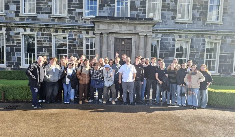 A group of students and staff pose for a photograph outside a historic stone building on a sunny day.