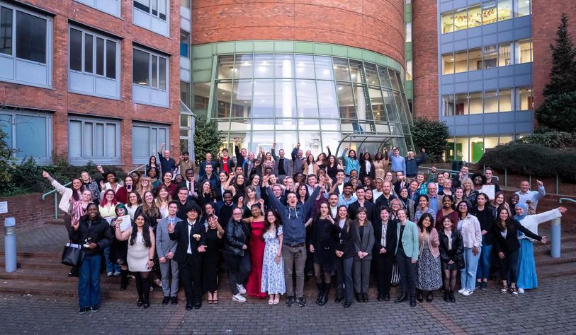 A large group of people cheering and smiling outside a red brick building