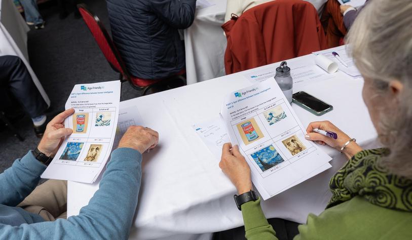 Several people sit around a table reviewing printed materials, with smartphones and papers on the table. Two people in the foreground hold illustrated information sheets.