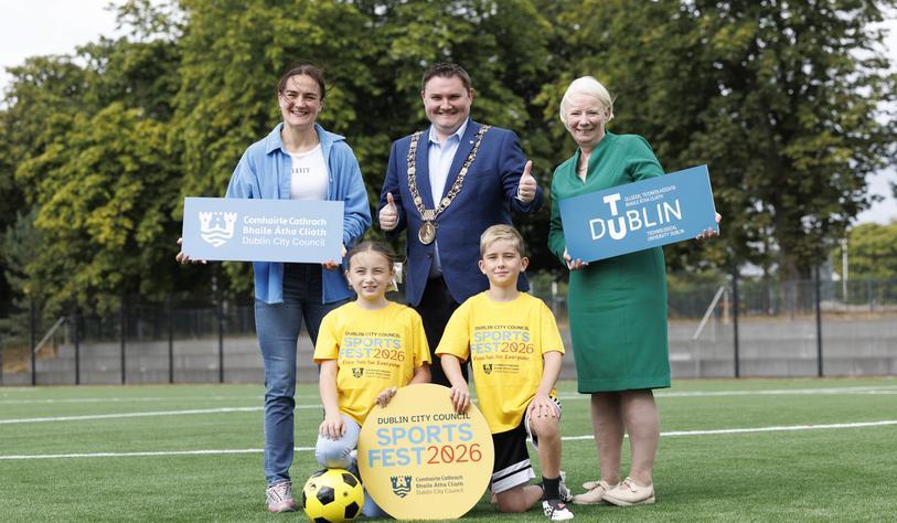 A group of four adults and two children pose on a sports field for Dublin City Council SportsFest 2026, holding TU Dublin and Dublin City Council signs, with a football and SportsFest branding in the foreground.