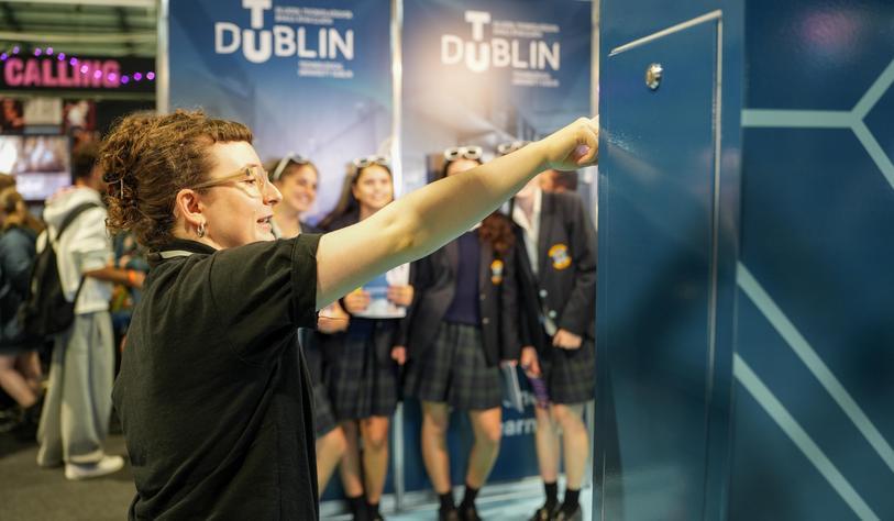 A person interacts with an exhibit at the TU Dublin stand during an education event, with TU Dublin banners and other students visible in the background.