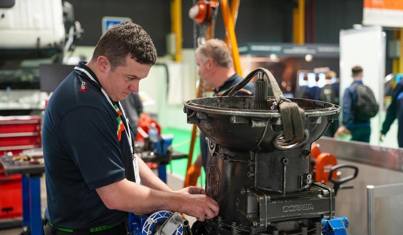 An apprentice in a workshop adjusts a large industrial machine while wearing a dark uniform and safety lanyard. Other workers and equipment are visible in the background.
