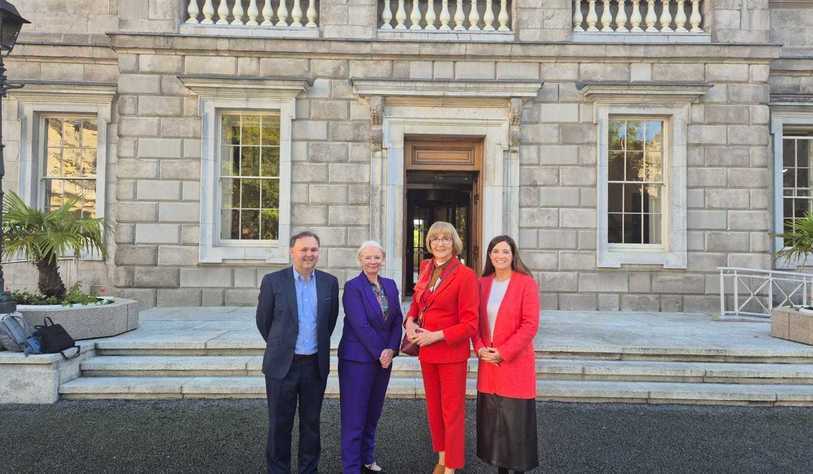 Four adults stand together outside a grand historic stone building, posing for a group photo. Three women wear colorful suits, while a man wears a dark suit with a blue shirt. The building features large sash windows, stone steps, decorative balustrades, and potted plants.