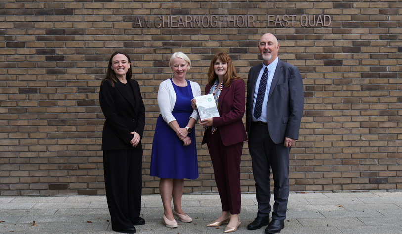 Four people stand outside a university building, with a brick wall and university signage behind them. One woman holds a certificate or document, while the others stand beside her smiling.