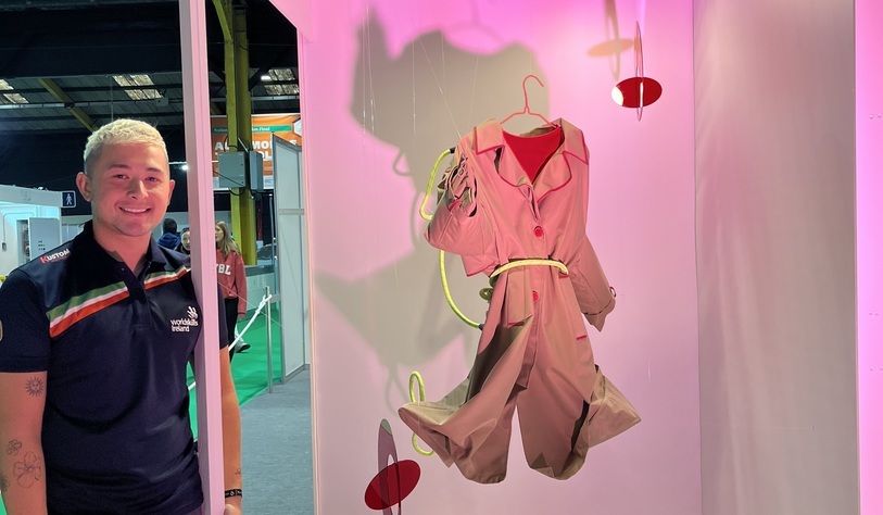 A student stands beside a fashion display featuring a pink and beige outfit suspended against a pink backdrop at an exhibition.