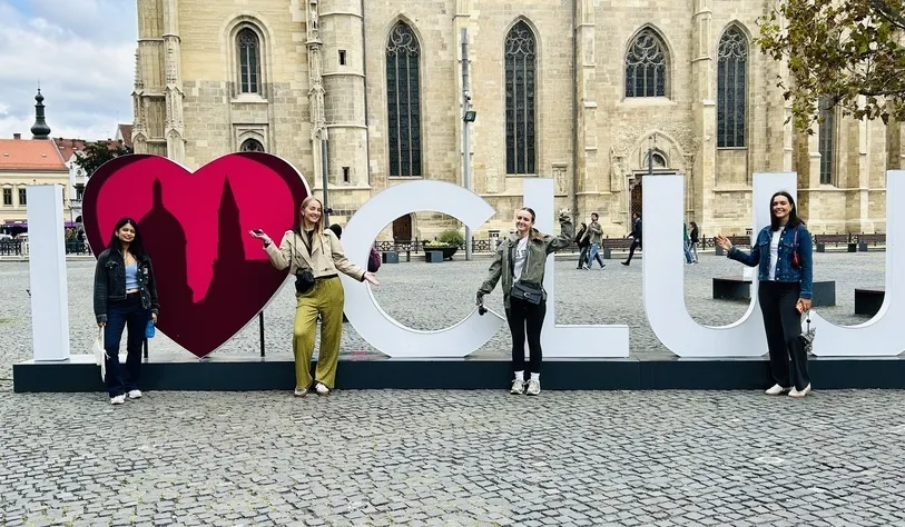 Four students pose beside a large “I ♥ CUL” sign in a cobblestoned European square, with a historic Gothic church visible in the background.