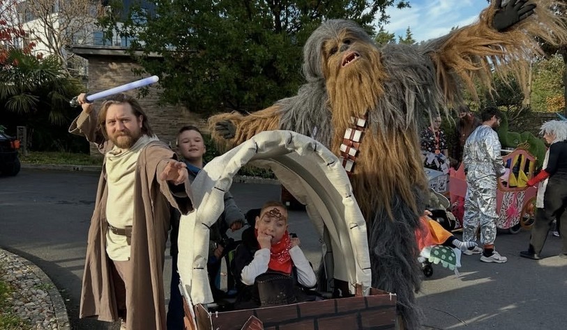 A person dressed as a Jedi, holding a lightsaber, stands alongside a child and a participant dressed as Chewbacca, with a homemade wooden-style cart in the foreground.