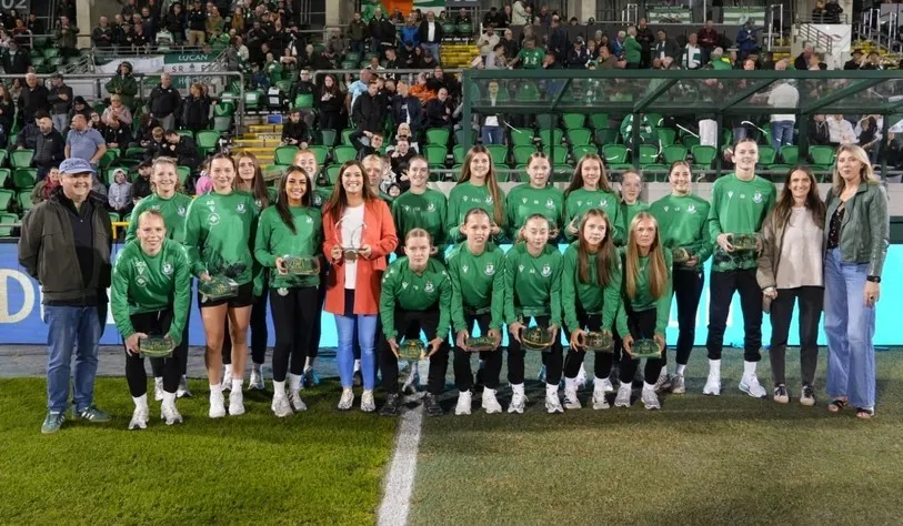 A women’s football team in green jerseys poses on the pitch with coaches and staff, holding trophies, in front of a crowded stadium stand with green seats.