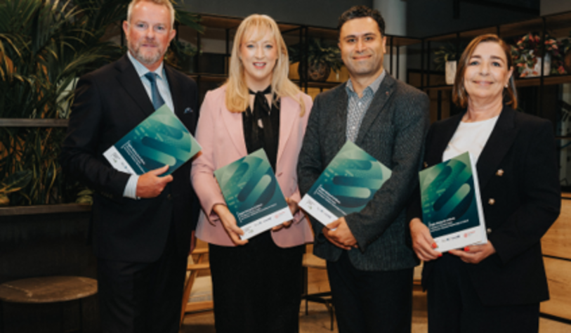 Four professionally dressed adults stand together indoors, each holding a teal-and-green booklet or award, posing for a group photo in a modern office with plants and wood furnishings.
