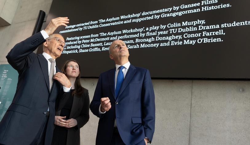 Three people in formal attire looking up at a large projected screen displaying information about a TU Dublin student theatre production.