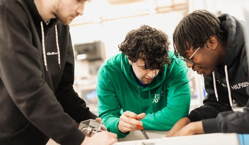Three students collaborate on a hands-on electronics project, leaning over a workbench and examining a circuit board and equipment together.