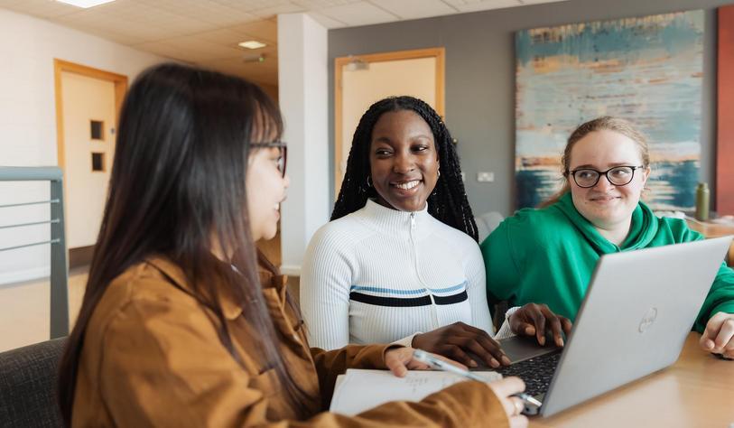 Three students collaborate around a laptop in a bright study space, smiling and discussing their work together.