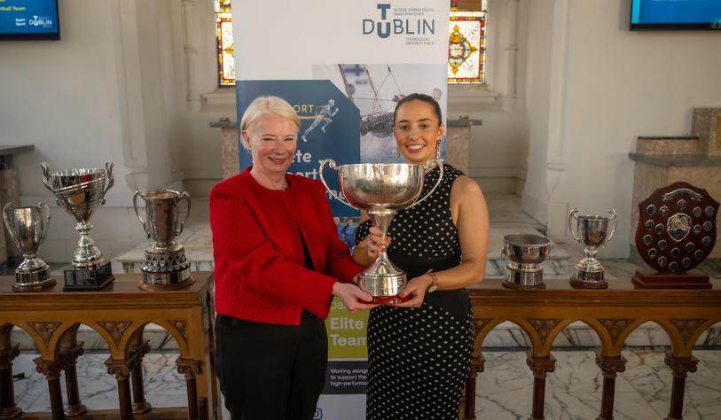A person hands a trophy to another person, both are smiling