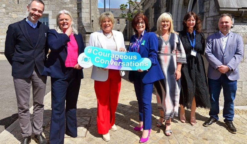 Seven adults standing outdoors in a cobblestone courtyard between stone buildings, posing together and holding a large turquoise sign that reads “#Courageous Conversations.” The group is dressed in business or smart-casual clothing, and the scene is lit by bright sunlight with architectural details and a tree visible in the background.