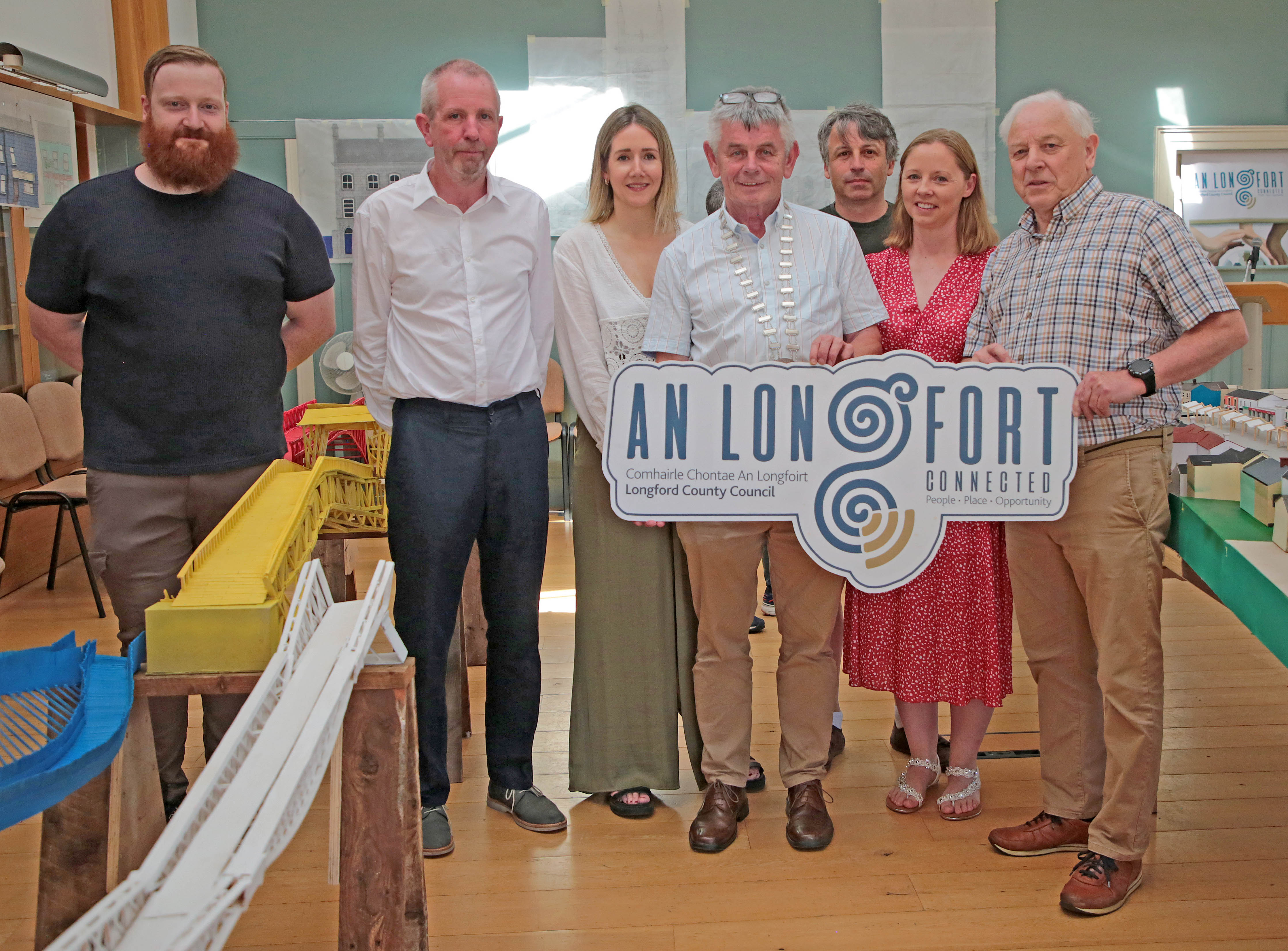 A group of seven people stand together indoors, posing for a photo while holding a sign that reads 
