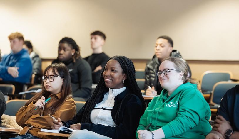 Students seated in a university lecture hall, attentively listening and smiling, with notebooks and pens in hand; the group reflects a diverse mix of backgrounds, and one student wears a green TU Dublin hoodie.