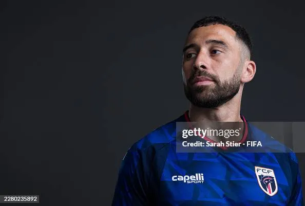 A man (Roberto Lopez) with a beard wearing a blue soccer jersey looks intently to the right. The dark background highlights his serious expression and focused demeanor.