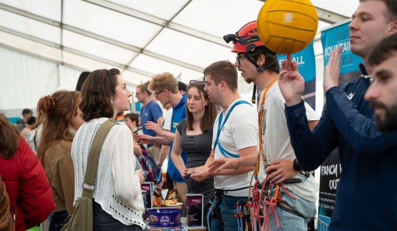 Busy indoor event with people chatting at a booth; a climber in a helmet holds an orange ball, creating a lively, friendly mood.