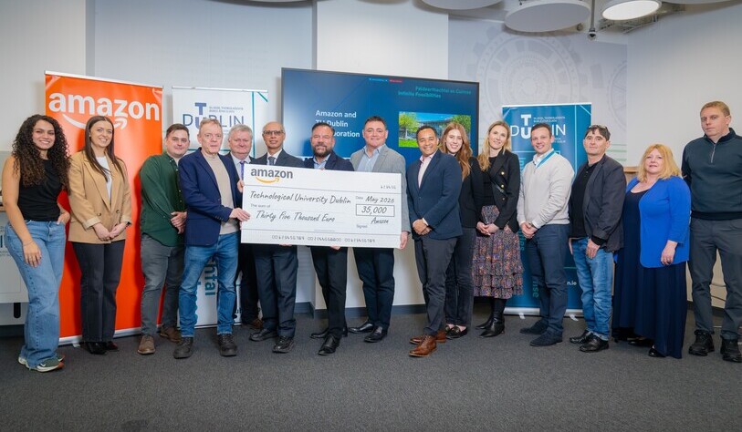 A group of people smiling and standing in a row indoors, holding a large check for thirty-five thousand euros from Amazon to Technological University Dublin. Various banners are visible in the background.