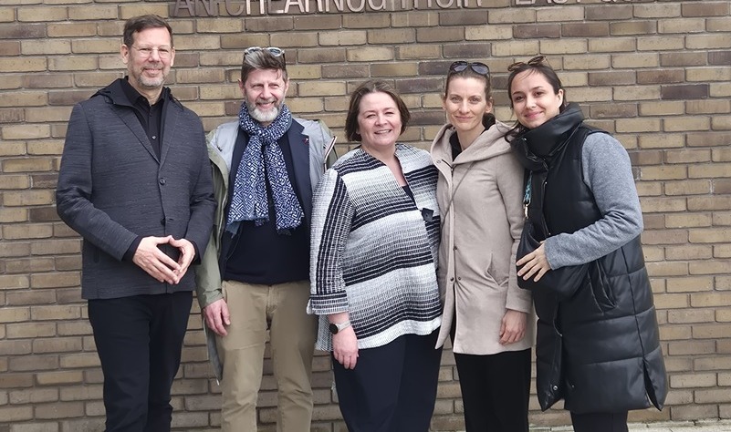 Five people stand closely together, smiling against a brick wall background. Their relaxed poses and warm smiles convey a sense of friendship and camaraderie.