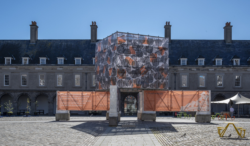 A large building with a cube-shaped structure covered in patterned fabric and orange mesh panels, set in a cobblestone courtyard under a clear blue sky.
