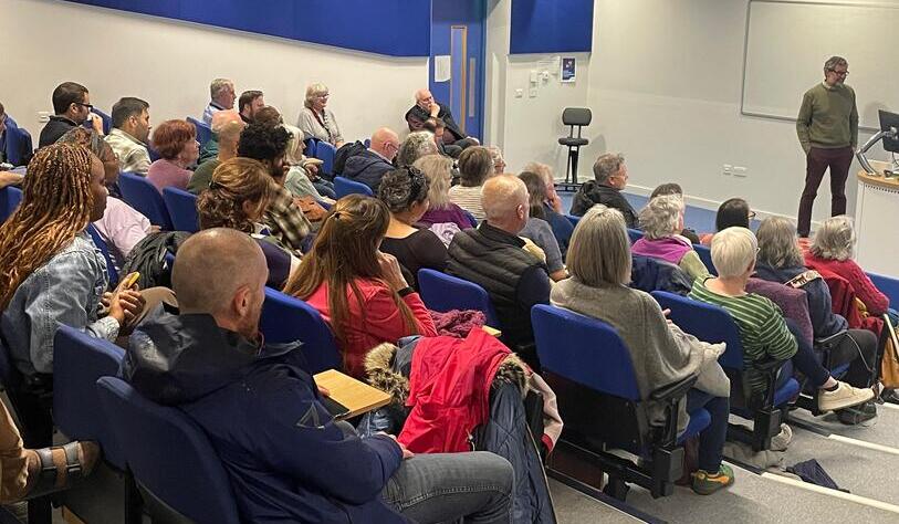 A speaker stands at the front of a lecture hall, engaging an attentive audience seated in blue chairs. The diverse group is focused and attentive.