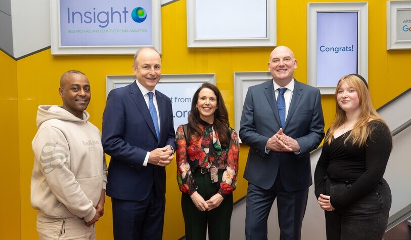 Five people stand smiling in front of a yellow wall with framed signs, including one that reads 