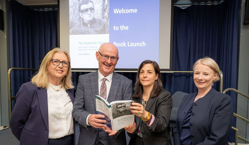 Four people stand smiling, holding a book, in front of a presentation screen at a book launch event. The atmosphere is formal and celebratory.