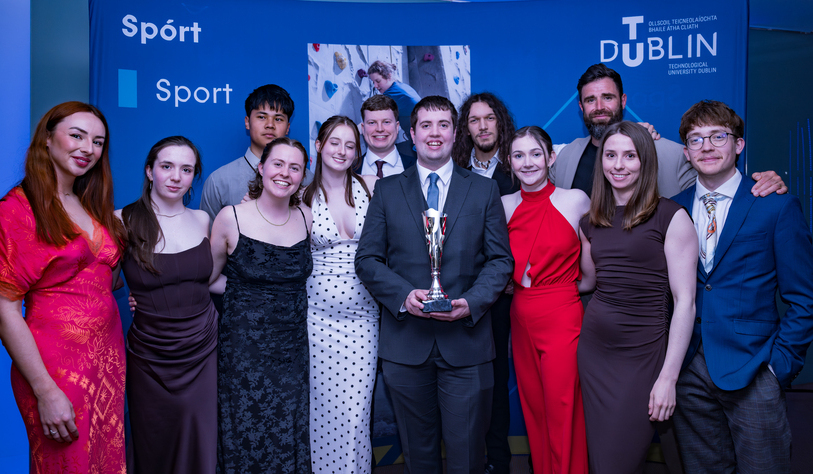 A group of eleven young adults dressed formally, smiling and posing with a trophy against a blue backdrop with 