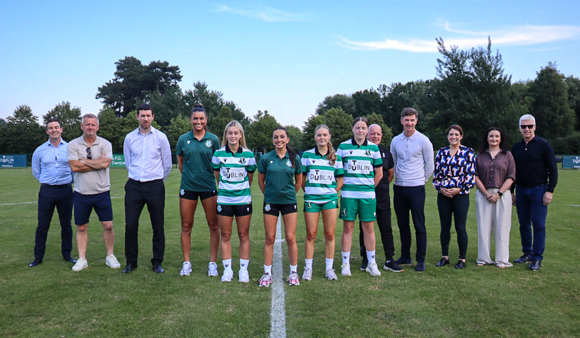 A group photo on a grassy field featuring five soccer players in green and white uniforms, flanked by six men and five women dressed in smart casual attire. A clear blue sky and trees are visible in the background, conveying a sense of teamwork and unity.